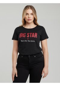 Fiatal nő fekete "BIG STAR Your Life. Your Jeans." feliratú pólóban és fekete farmerral, egyszínű szürke háttér előtt áll.
