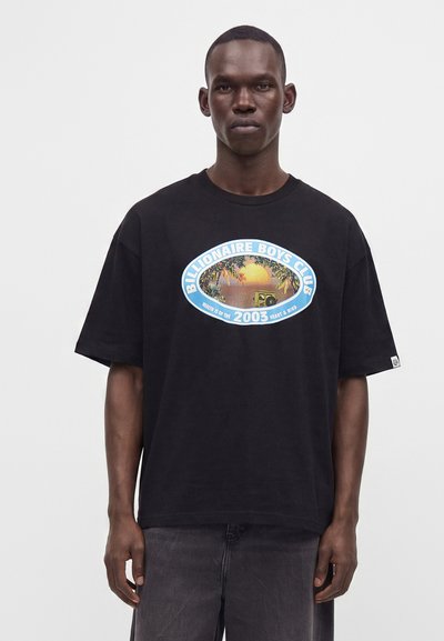 Man wearing a black oversized T-shirt with a colorful "Billionaire Boys Club" logo featuring a sunset, trees, and a car.