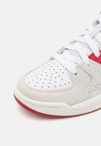 Záběr na špičku bílo-beige tenisky s perforovanou kůží, bílými tkaničkami, červeným akcentem, semišovými detaily a červeným okrajem podešve.