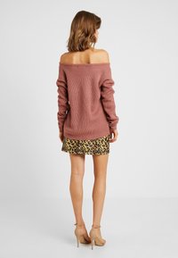 Femme portant un pull mauve à épaules dénudées, une mini-jupe imprimé léopard et des sandales à talons hauts couleur nude, debout, de dos, sur fond blanc.