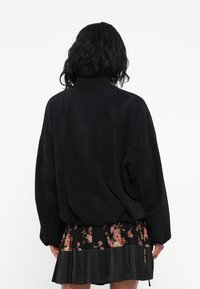 Svart cordjakke med høy krage, oversized passform og senkede skuldre. Kombinert med et plisserte blomsterkjole for kontrast.