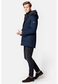 Granatowa parka z kapturem, z zamek błyskawicznym z przodu, bocznymi kieszeniami i teksturowaną podszewką. W zestawie z ciemnymi dżinsami i brązowymi butami.