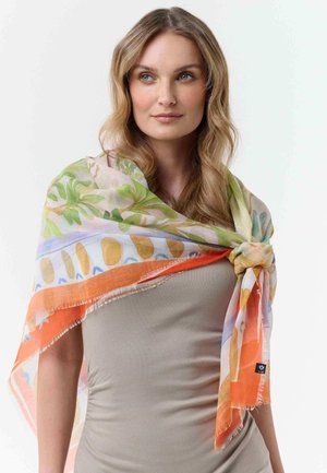 Frau mit langen blonden Haaren, die ein beigefarbenes Oberteil und einen bunten, floralen Schal trägt, der locker über ihre Schultern gelegt ist, vor weißem Hintergrund.