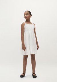Weißes Kleid mit Spitze, kreisförmigem Schnitt und bestickten Blumemustern. Kombiniert mit flachen schwarzen Schuhen. Einfaches, fließendes Design.