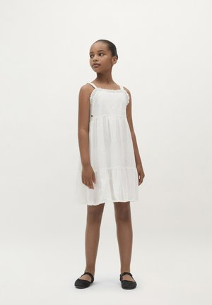Weißes Kleid mit Spitze, kreisförmigem Schnitt und bestickten Blumemustern. Kombiniert mit flachen schwarzen Schuhen. Einfaches, fließendes Design.