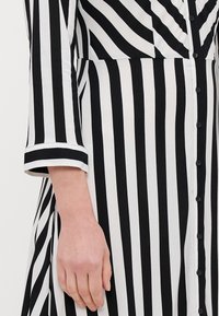 Dłoń oparta z boku, ubrana w czarno-białą pionową w paski koszulę z guzikami i rękawami do trzech czwarte. 