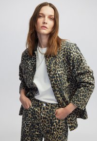 Giacca con stampa leopardata e pantaloni coordinati in denim beige e nero, con un design con colletto e tasche frontali. Modello che indossa una T-shirt bianca.