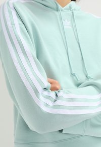 Vihreä huppari, jossa on valkoiset kolmen raidan korostukset hihoissa, kengurun tasku, resorit hihansuissa ja säädettävä nyörillä varustettu huppu.