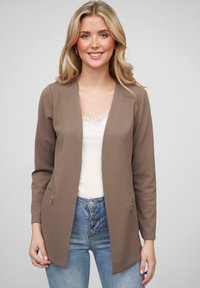 Cardigan brun texturé avec une coupe décontractée, devant ouvert et poches latérales zippées, porté sur un haut clair et un jean bleu.