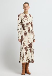 Długa sukienka w kwiaty z brązowym i kremowym wzorem, wykonana z elastycznego materiału, zestawiona z brązowymi, skórzanymi butami do kolan.