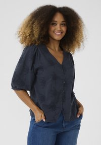 Blusa navy con scollo a V, maniche corte a sbuffo e intricato ricamo floreale. Presenta bottoni lungo la parte anteriore e una vestibilità comoda.