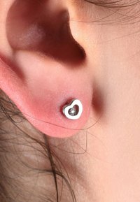 Silberne, herzförmige Steckohrringe mit einem klaren Edelstein in der Mitte. Glatte, glänzende Oberfläche mit einem schlichten Design, auf einem warmen Hautton präsentiert.