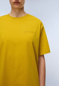 Personne portant une chemise à manches courtes jaune moutarde avec un petit logo en relief "NAPAPIJRI" sur la poitrine gauche devant un fond clair.