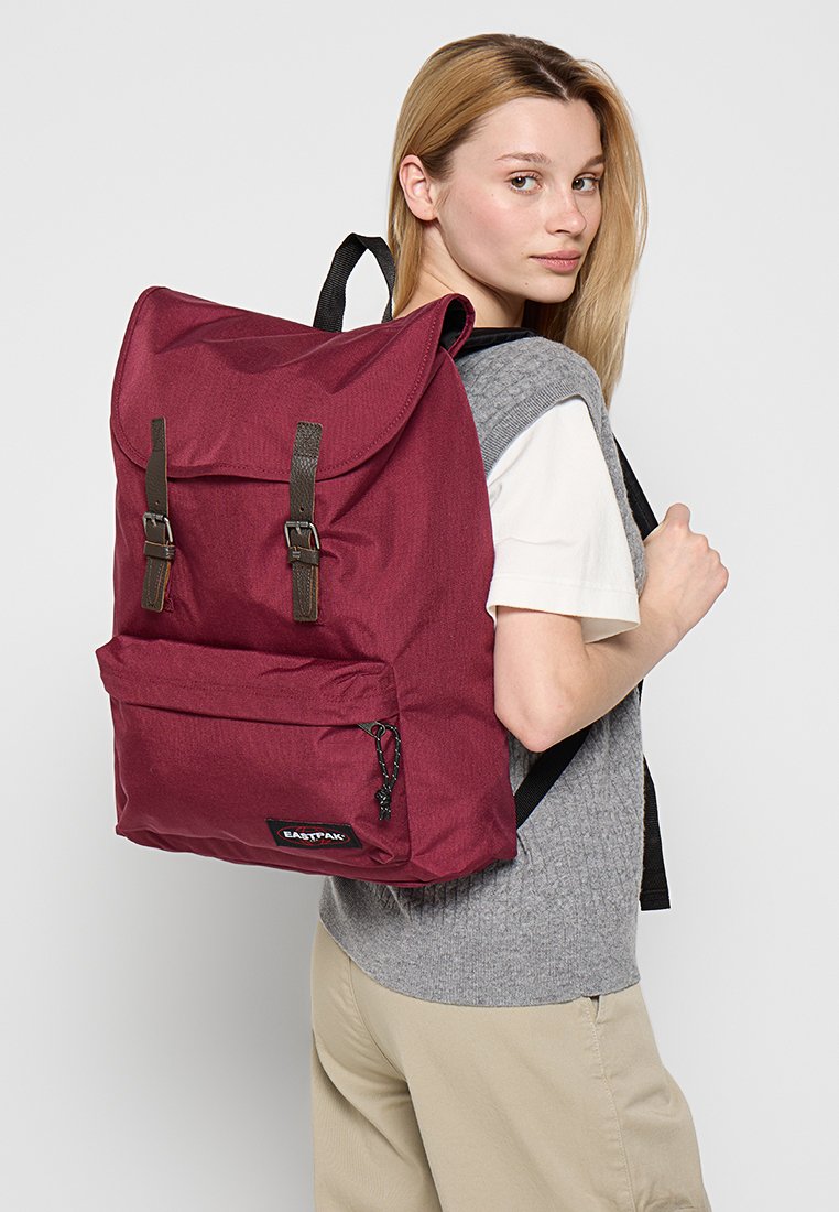 Bordeauxfarbener Stoff-Rucksack mit einem Rollverschluss, zwei braunen Riemen und einer Fronttasche. Mit einem Markenlogo an der Unterseite vorne. Leichtes Design.