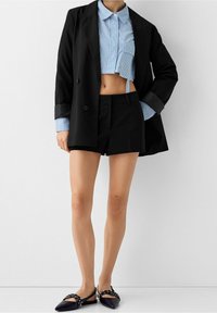 Žena v crop topu s modrými proužky, černém dvouřadém saku, černých šortkách a černých špičatých plochých botách se sponami a nýty.