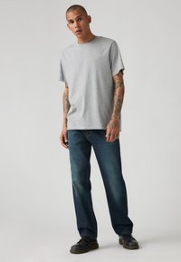 Grijze katoenen t-shirt met een ronde hals, gecombineerd met donkerblauwe spijkerbroek. De spijkerbroek heeft een recht gesneden pijp en lichte vervaging. Zwarte schoenen.