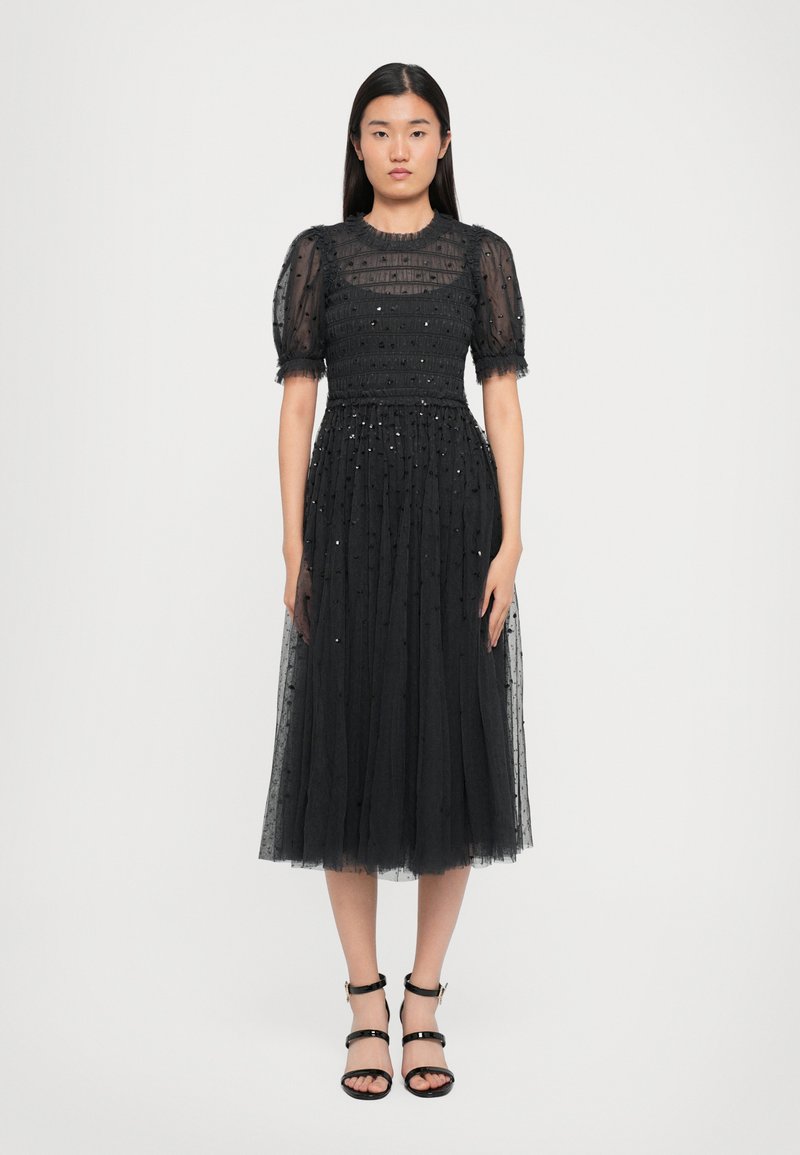 Zwart tule jurk met korte puffmouwen, textuur op het lijfje, elastische taille en verspreide versieringen. Gepaard met zwarte sandalen met banden.