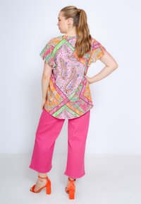 Bunte gemusterte Bluse mit einer Mischung aus pinken, orangen und grünen Paisleymustern, kombiniert mit leuchtend pinken, weiten Hosen und orangen Absätzen.