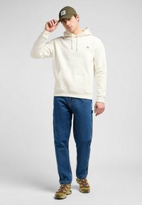 Felpa bianca con tasca frontale e cordino, abbinata a jeans blu scuro. Il modello indossa un cappellino verde oliva e sneakers marroni con accenti blu.