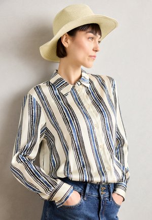 Blouse rayée en crème, marine et noir, avec un devant à boutons, des manches longues et une texture douce et lisse. Associée à un jean en denim.
