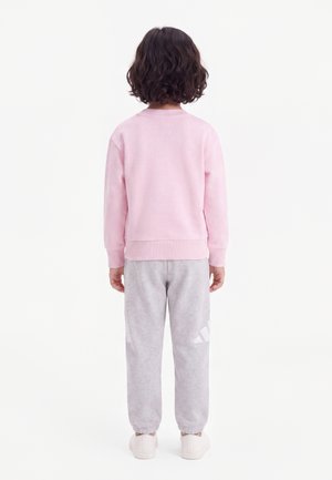Enfant aux cheveux courts, bouclés et foncés, portant un sweat-shirt rose clair, un pantalon de jogging gris et des baskets blanches, debout de dos sur fond blanc.