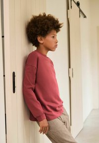 Punane pikkade käistega pullover, ümmarguse kaelusega ja vabalt istuv; koos ruuduliste pükstega. Taustal on neutraalne sein.