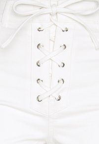 Pantalones cortos de mezclilla blanca con un cierre frontal tipo lazo con ojales, atados con cordones blancos. La tela tiene una apariencia suave y texturizada.