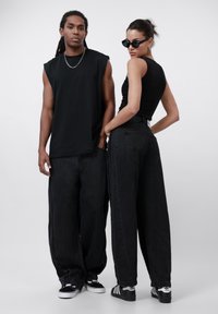 Black sleeveless top paired with wide-leg black denim jeans. Model in sunglasses and chain necklace, showcasing a relaxed streetwear style.