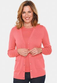 Koraalrode cardigan met een geribbelde textuur, v-hals, knoopsluiting en twee voorzakken. Heeft lange mouwen en een zachte stof.