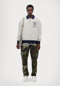 Šedá mikina s námorníckym golierom, zipsom vpredu a logom. Camo cargo nohavice s viacerými vreckami. Biele tenisky, taška na ramene.