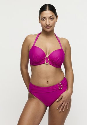 Vrouw met donker haar in een magenta bikini met gouden gespsdetails, staand tegen een eenvoudige lichte achtergrond.