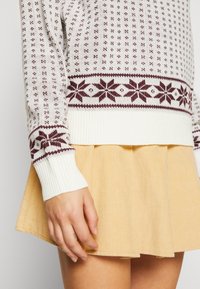 Pull en maille blanc avec des motifs géométriques bordeaux, associé à une jupe courte en velours côtelé jaune texturé. Détails côtes aux poignets et à l'ourlet.
