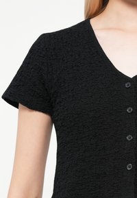 Haut court à manches courtes noir texturé avec un décolleté en V et six boutons noirs sur le devant. Le matériau semble froissé avec une surface douce.