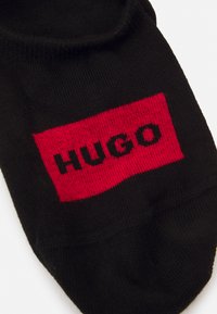 Čierna ponožka s červeným obdĺžnikovým nášivom, na ktorom je v tučných čiernych písmenách napísané "HUGO". Hladký povrch s rebrovaným otvorom na členku.