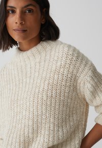 Maglione spesso di crema con collo alto, texture a coste e vestibilità ampia. Presenta una morbida stoffa lavorata a maglia con sottili variazioni di tonalità.