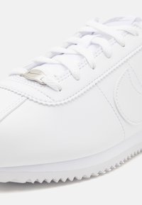 Gros plan d'une basket en cuir blanche avec des lacets blancs, une étiquette métallique sur les lacets, des coutures détaillées et une semelle en caoutchouc blanche rainurée.
