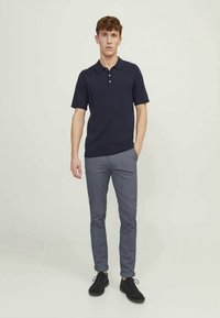 Young man standing straight wearing a navy polo shirt, gray slim-fit trousers, and black lace-up shoes against a plain background.