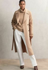 Beige wollen jas met een dubbelzijdig ontwerp over een beige coltrui, gecombineerd met witte broek en bruine enkel laarzen. Gladde texturen.