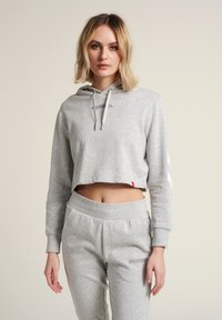 Sweat à capuche court gris avec cordons, portant le logo "HUMMEL" sur la poitrine, accompagné d'un pantalon de survêtement gris assorti et de subtils accents sur les côtés.