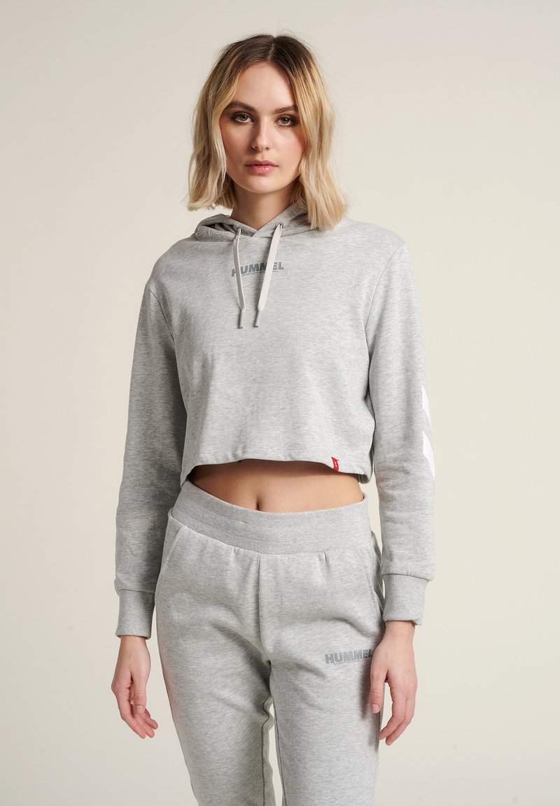 Sweat à capuche court gris avec cordons, portant le logo "HUMMEL" sur la poitrine, accompagné d'un pantalon de survêtement gris assorti et de subtils accents sur les côtés.