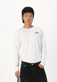 White long-sleeve shirt with a round neckline and a small black logo on the chest. Paired with black denim jeans. Simple and unembellished design.