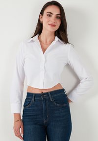 Witte, geknipte button-up blouse met een kraag, lange mouwen en een gladde textuur, gecombineerd met donkerblauwe goed passende jeans.