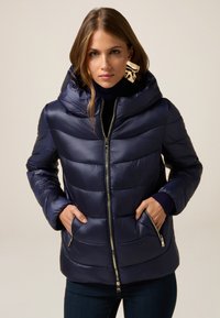 Marineblå pufferjakke med quiltet design, oversized hætte, lynlåslukning foran og sidelommer; lavet af glansfuldt, glat stof.