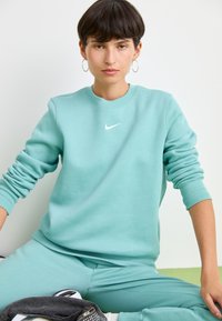 Lichtblauw sweatshirt met ronde hals en omgeslagen mouwen, met een klein wit Nike-logo op de borst. Bijpassende teal joggingbroek.