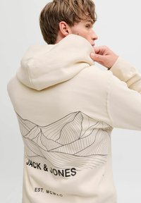 Hoodie écru avec un dessin en ligne noire de montagnes au dos, avec "JACK & JONES" et "EST. MCMXC" imprimés en dessous.