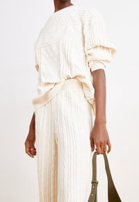 Krämfärgad kabelstickad tröja och matchande byxor med ribbade muddar och midja. Håller en grön läderremm väska. Strukturerat, mjukt material.