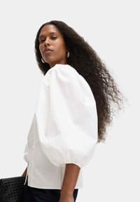 Witte blouse met pofmouwen van lichte stof, met knoopsluiting en een relaxed fit, gecombineerd met een zwarte gehaakte handtas.