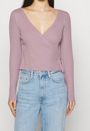 Kobieta w bluzce z długim rękawem w kolorze wrzosowym z koronkową lamówką, wiązaną z boku, w połączeniu z jasnoniebieskimi jeansami z wysokim stanem.