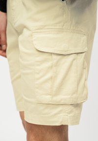 ΜπεζCargo σορτς με μεγάλο πλαϊνό καπάκι τσέπης, φορεμένα από άτομο με ορατή τρίχα στα χέρια και στα πόδια, πάνω σε απλό φόντο.