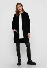 Black long coat with a straight cut, made from textured fabric. Features side pockets and is worn over a white top and black leggings.